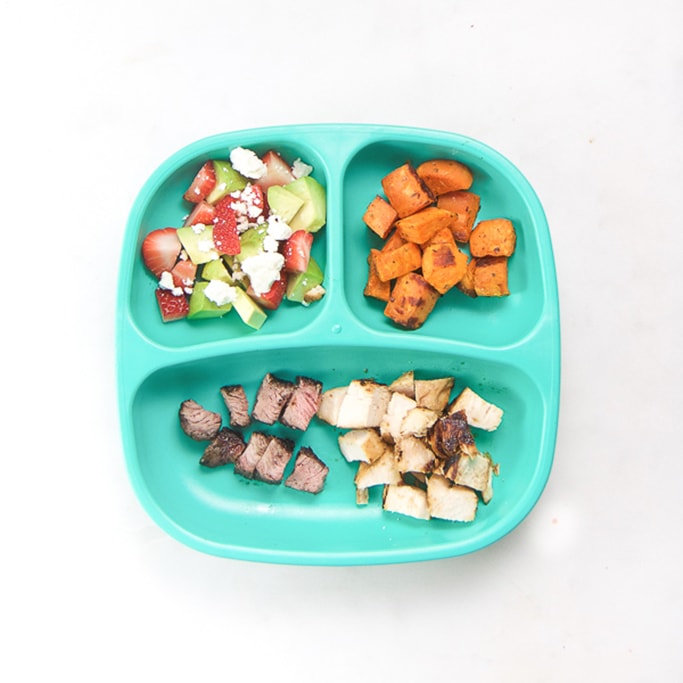 3-section plate on a white surface filled with toddlers dinner meal - cut up steak and chicken, roasted sweet potatoes, strawberries and avocado.