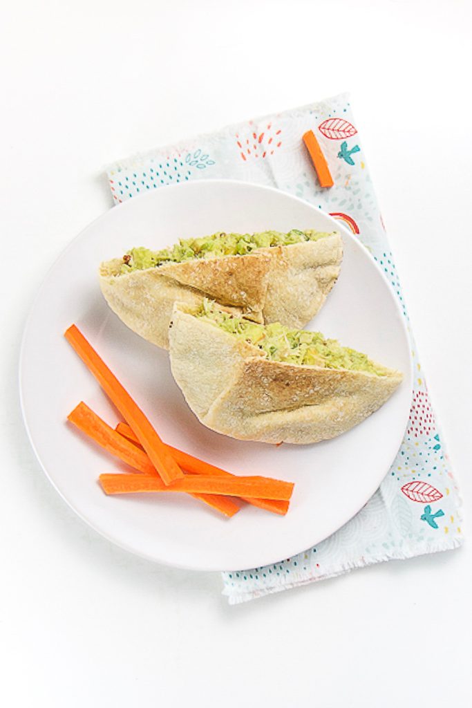 a white round plate is filled with a mini pita pocket cut in half and filled with an avocado tuna salad, there are cut sticks of carrots on the plate as well. The plate is sitting on a multi-colored napkin on a white surface.