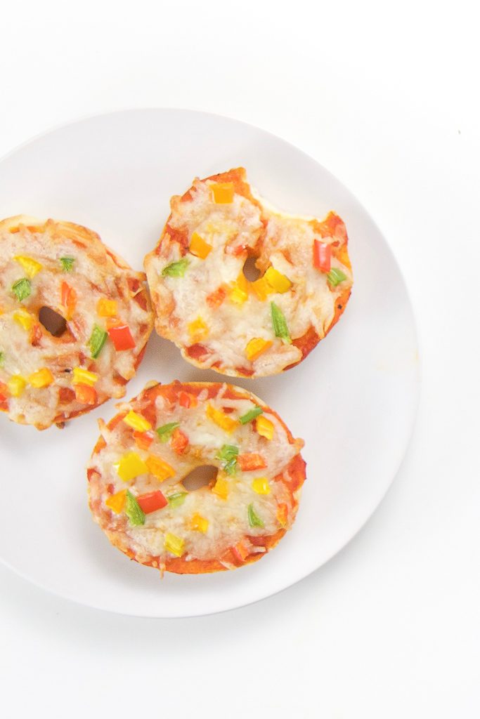 round white plate sitting on white surface. On plate are 3 mini bagels with pepper sprinkles, one bagel pizza has a bite taken out of it.