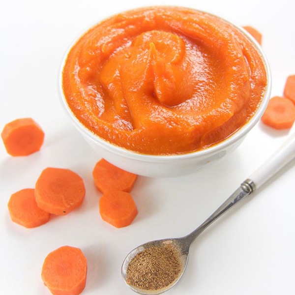 A small white bowl filled with carrot baby food puree with carrots and a spoon filled with nutmeg scattered around the bowl