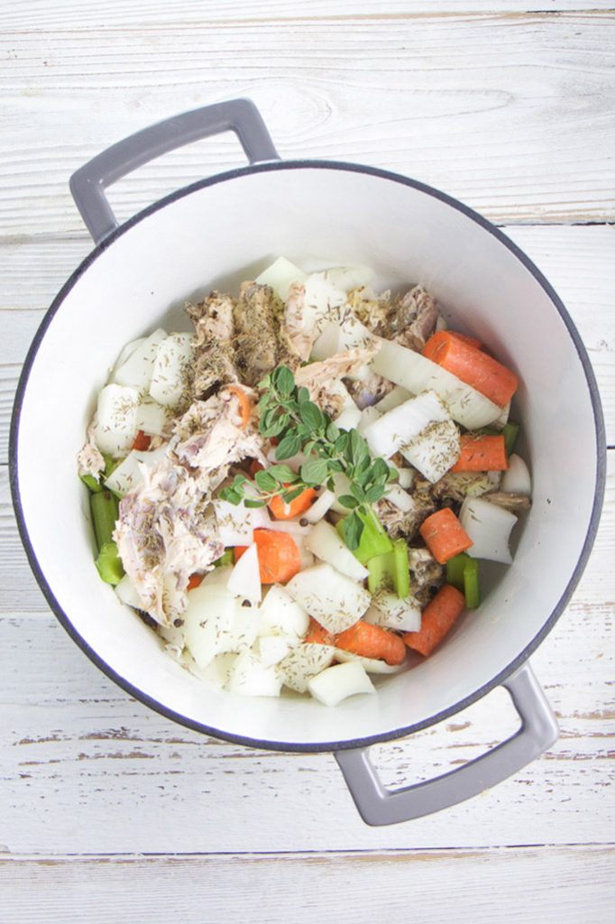 A white stockpot with grey rim on a wooden board filled with chicken bones, herbs and veggies.