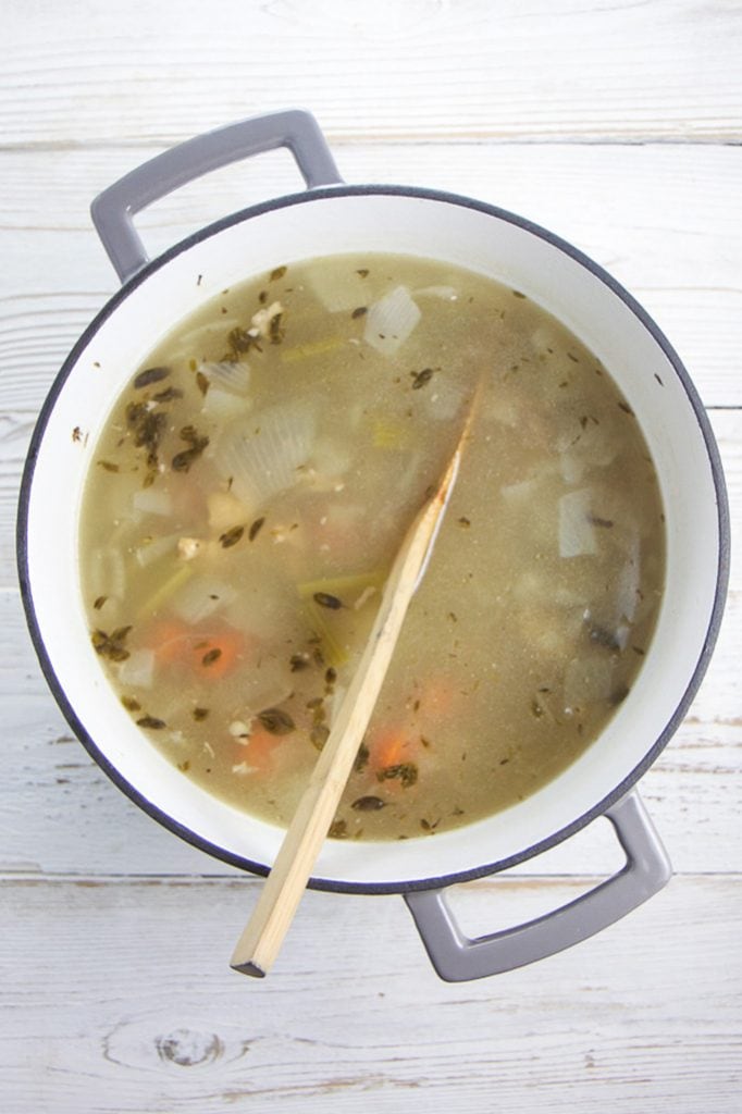A white stockpot with grey rim on a wooden board filled with chicken bones, herbs and veggies and water and simmered.