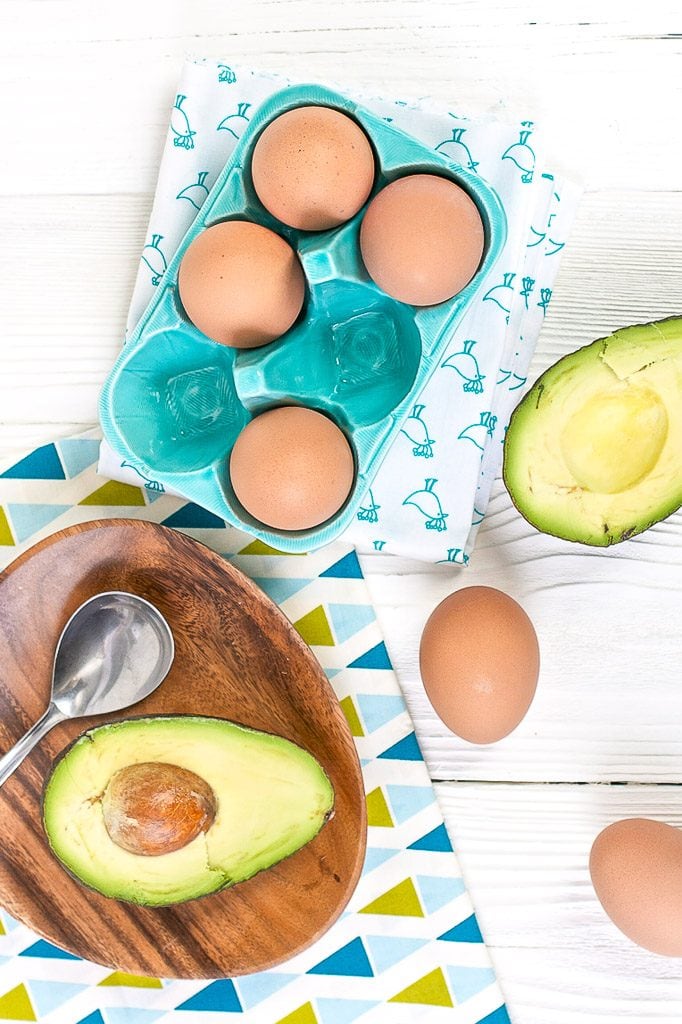Brown eggs and cut in half avocados are scattered about a white wooden board with napkins in teal and green.