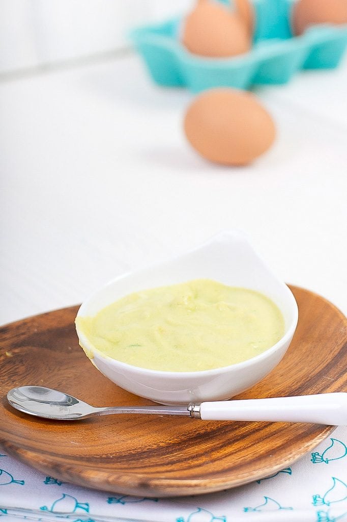 small white bowl is filled with an avocado and egg baby food puree, in the background are some brown eggs scattered around.