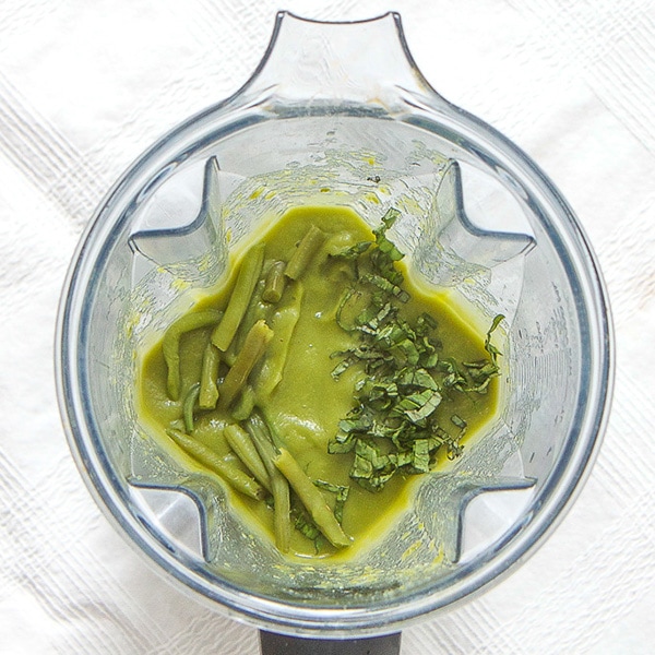 A clear blender is filled with a green bean puree with chunks of green beans and parsley on top