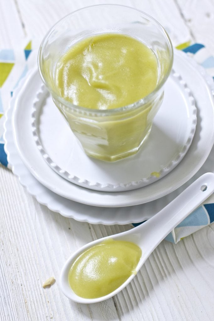 A clear glass jar is sitting on top of 2 white round plates. There is white spoon sitting next to the jar and both the jar and spoon are filled with a leek baby food puree.