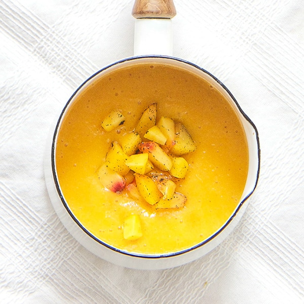 A small white saucepan is filled with a peach puree with chunks of peaches and vanilla beans sprinkled on top