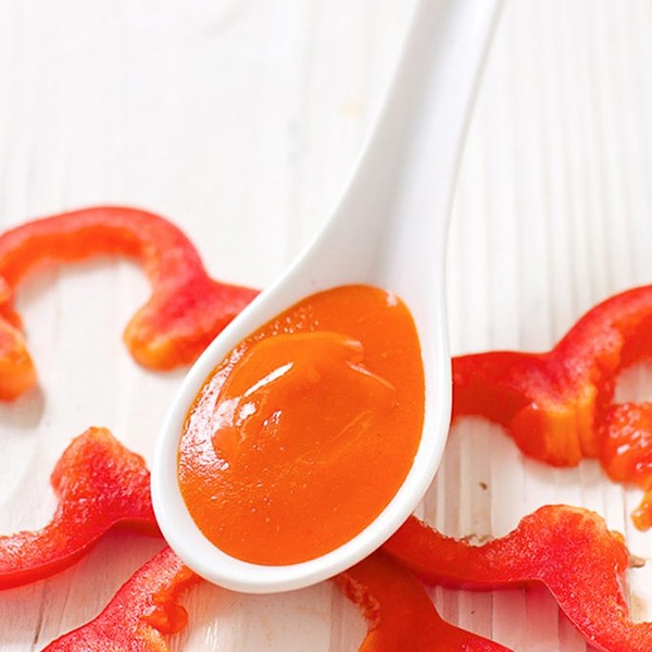 A white spoon is sitting on top of some cut red peppers with a red pepper puree inside of the spoon.