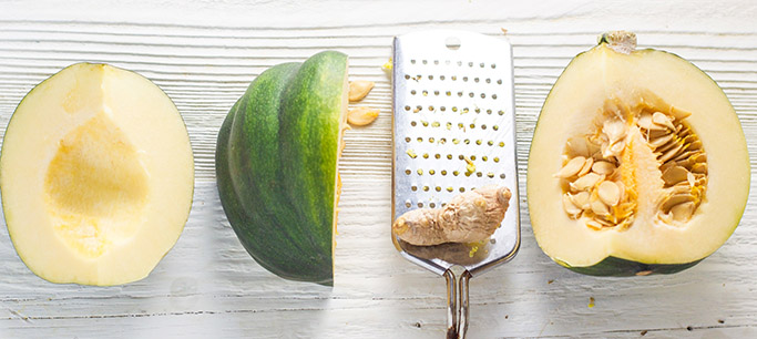 an acorn squash is cut open and in 3 pieces, some with seeds and one without. There is a piece of fresh ginger sitting on a mincing tool.