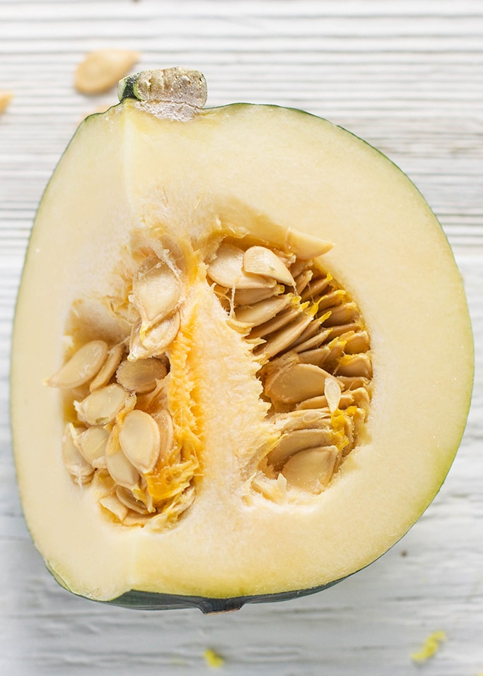 An up close of a cut open acorn squash with it's seeds still intact. The squash is sitting on a white wooden surface.