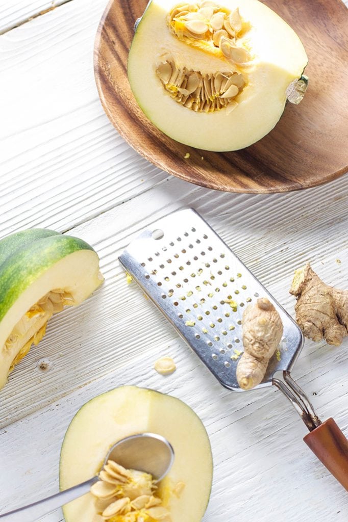 acorn squash is cut open and scattered about a white wooden surface. There is ginger root being minced as well.