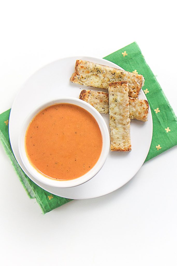 small white bowl filled with an easy creamy tomato soup is sitting on a white plate with 3 cheesy dippers next to the bowl. The bowl is on a green napkin sitting on a white surface.