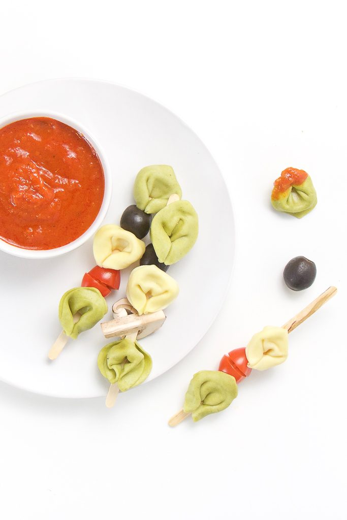 small white plate sitting on a white surface. On top of plate is a small white bowl filled with a dipping sauce and 3 wooden sticks filled with tortellini and veggies, a couple of tortellini and veggies are scattered on the table.