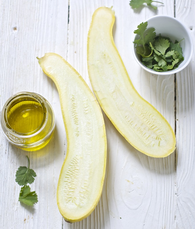 One cut open yellow squash laying on a white wooden board with a little jar of olive oil and a bowl of fresh cilantro next to it.