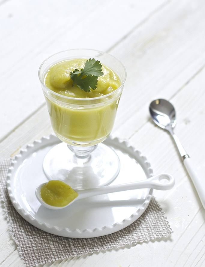 A clear jar filled wit a creamy yellow squash baby food puree. The jar is sitting on a white plate on a white wooden surface. 