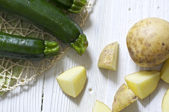 A group of 3 zucchini in the corner of the frame with a few scattered potatoes in the other, both sitting on a white wood surface.