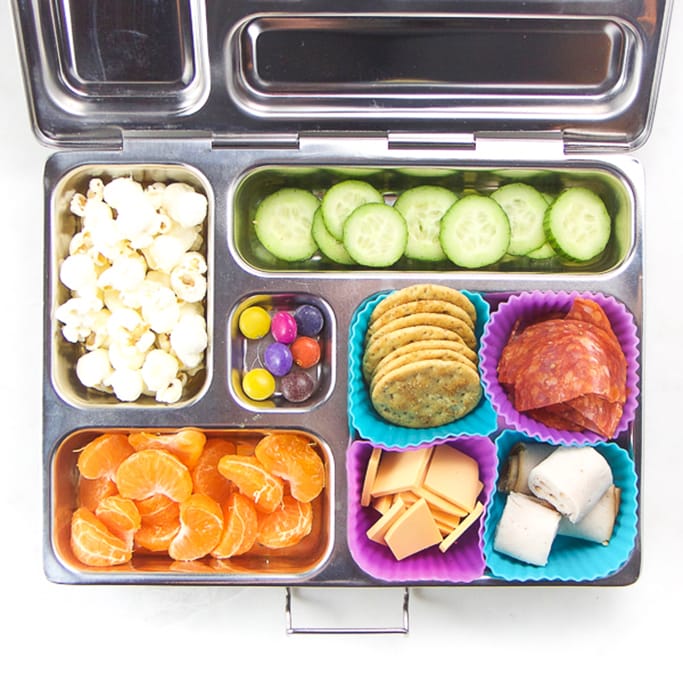 silver planetbox filled with crackers, cheese, turkey, salami, cucumbers, popcorn and orange slices.