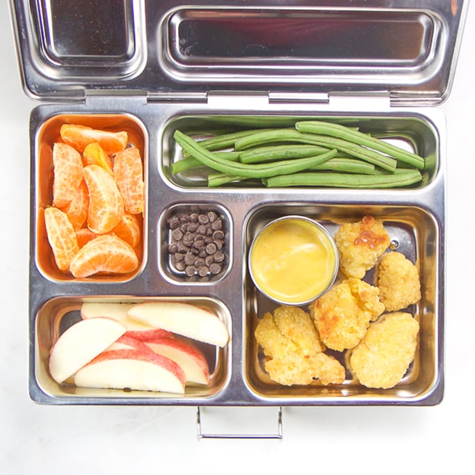 lunch box for kids filled with gluten free chicken nuggets, apple slices, green beans, oranges and chocolate chips