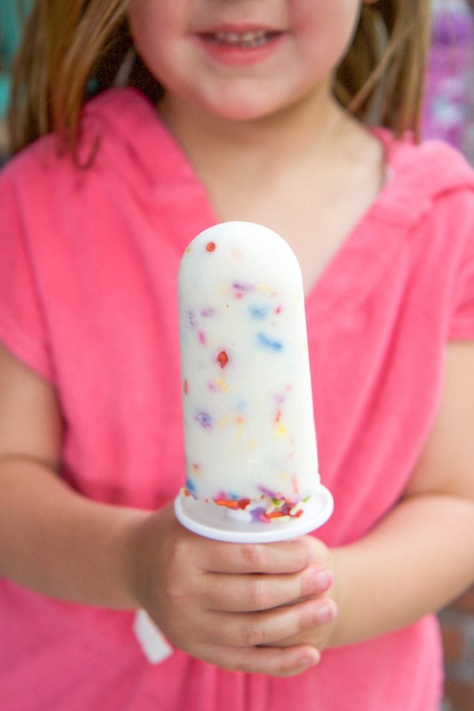 A small girl wearing pink holding a healthy funfetti popsicle out in front of her.