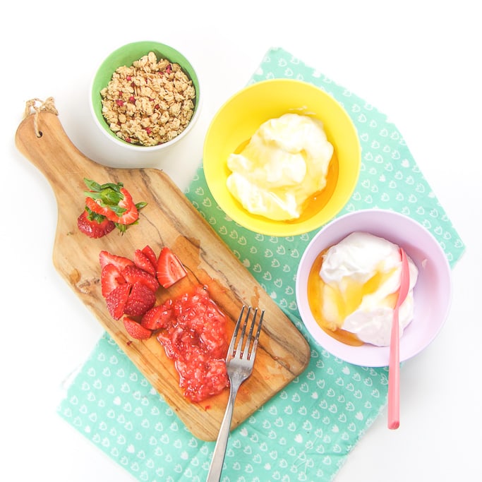 Bowls of yogurt, cutting board with smashed strawberries and granola - everything you need to make heathy yogurt parfaits for toddler and kids.