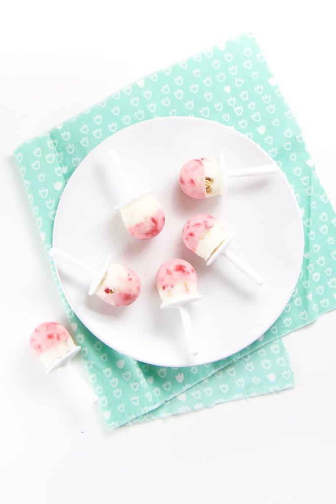 A white round plate with 5 healthy yogurt parfaits randomly on it, with one popsicle on the counter on a teal napkin.