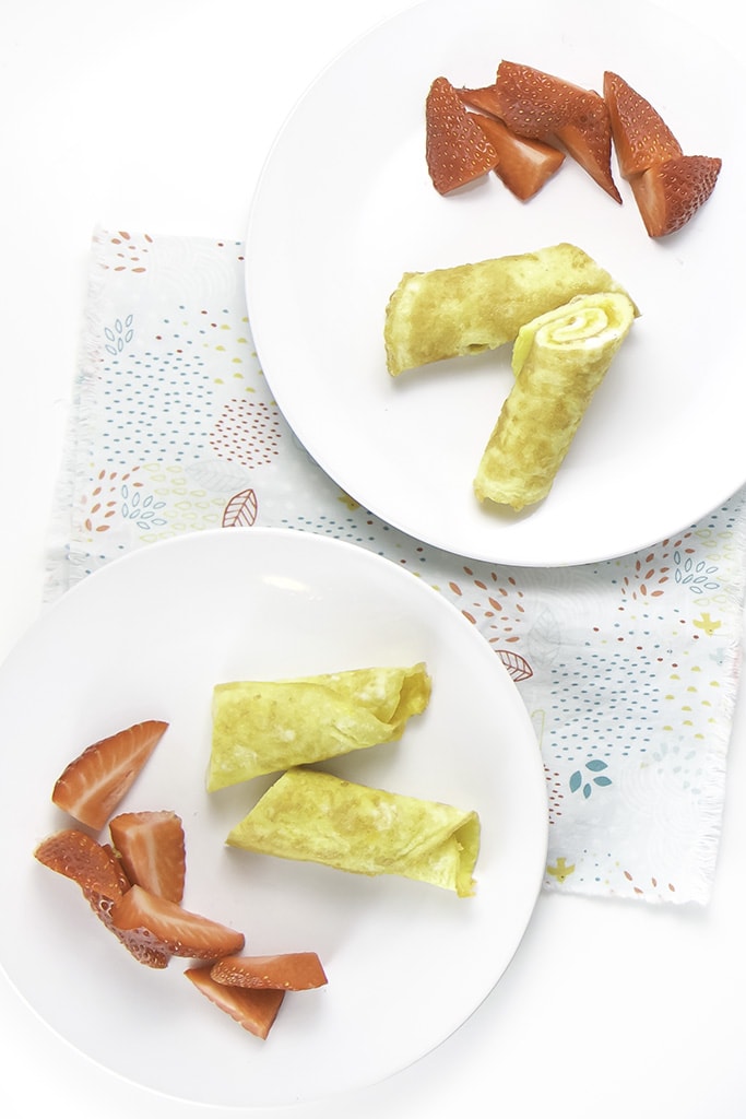 2 white round plates filled with an egg roll-up and strawberries, sitting on a multi colored napkin.