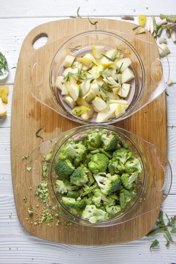 A cutting board with 2 steamer trays filled with broccoli, asparagus and beans.