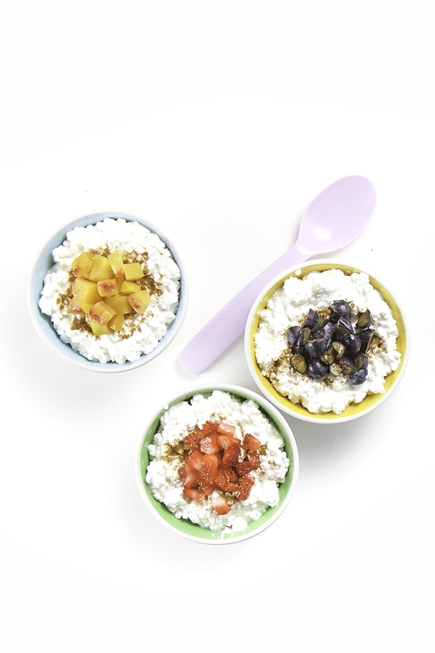 3 small bowls filled with cottage cheese an topped with chunks of fruit are lined up with spoons resting next to each bowl.