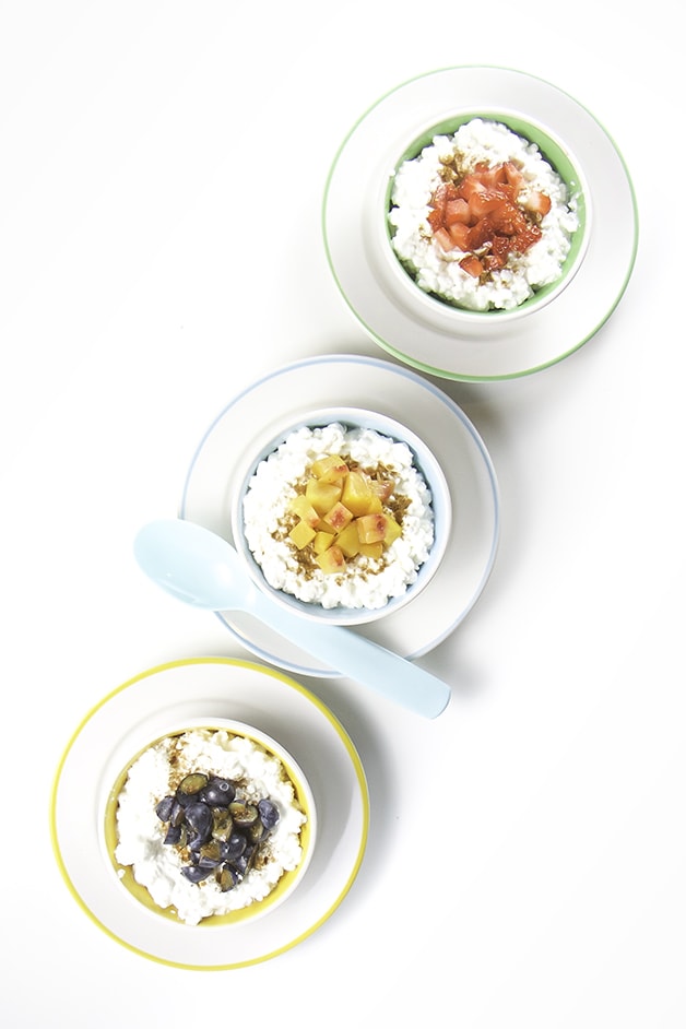 3 small bowls filled with cottage cheese an topped with chunks of fruit are lined up with spoons resting next to each bowl.