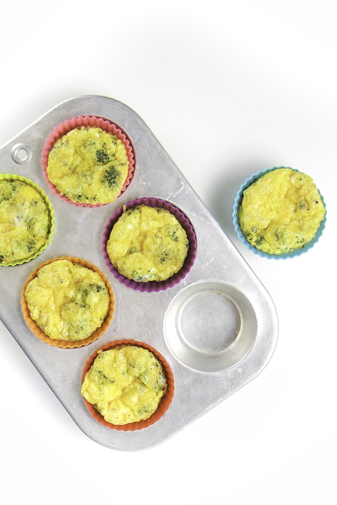 A silver muffin tin filled with broccoli egg cups with one cup on the white surface.