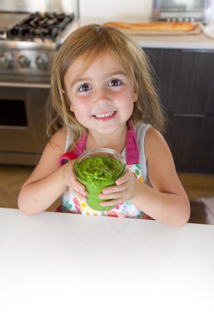 little kid in a kitchen, holding a jar of spinach basil pesto.