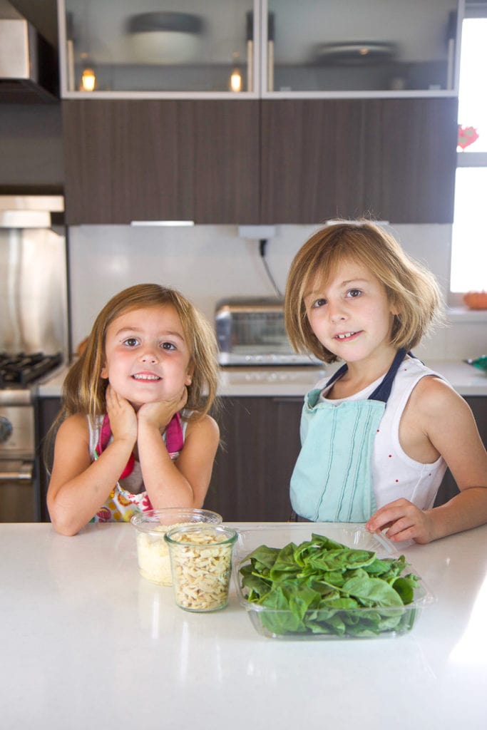 2 girls standing in a kitchen counter with a spread of recipe ingredients in front of them.