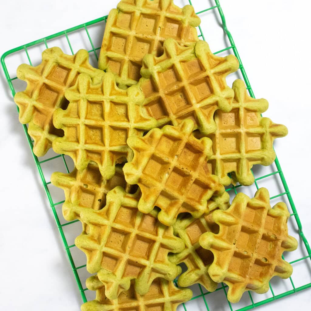 Green wire cooling rack on my kitchen counter with a bunch of spinach waffles cooling on top.
