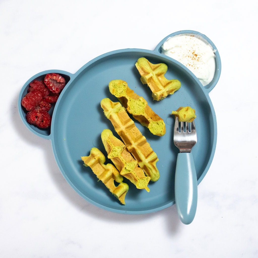 Blue kids plate on my white counter with strips of spinach waffles, cut raspberries and yogurt.
