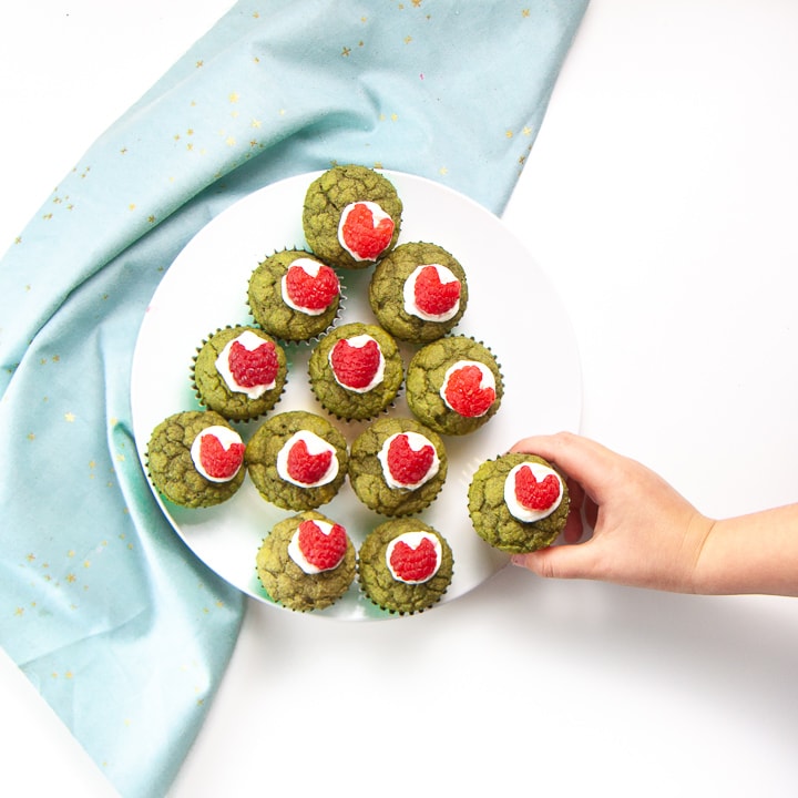 Green Grinch Muffins on a white plate with a small toddler hand reaching out to grab one.