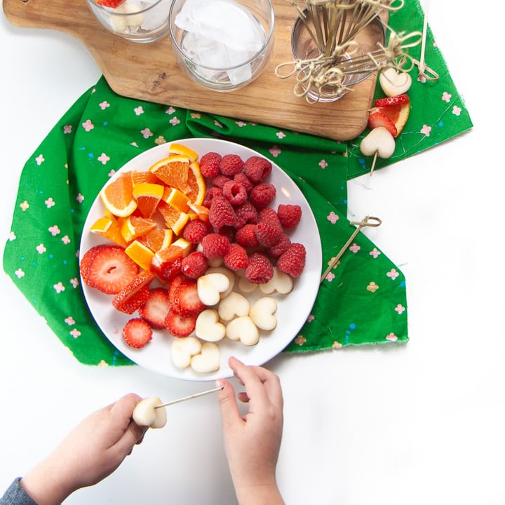 Round white plate with assortment of fruit on it with small kids hands making their own fruit sticks.