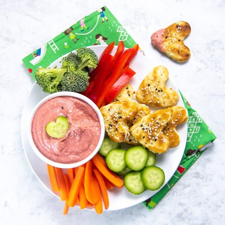 bowl of beet hummus with plate full of chopped veggies and cut-out heart pita bread.