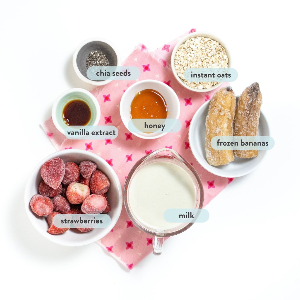 Ingredients spread on a white table with a pink napkin and labeled by name for a strawberry banana smoothie.