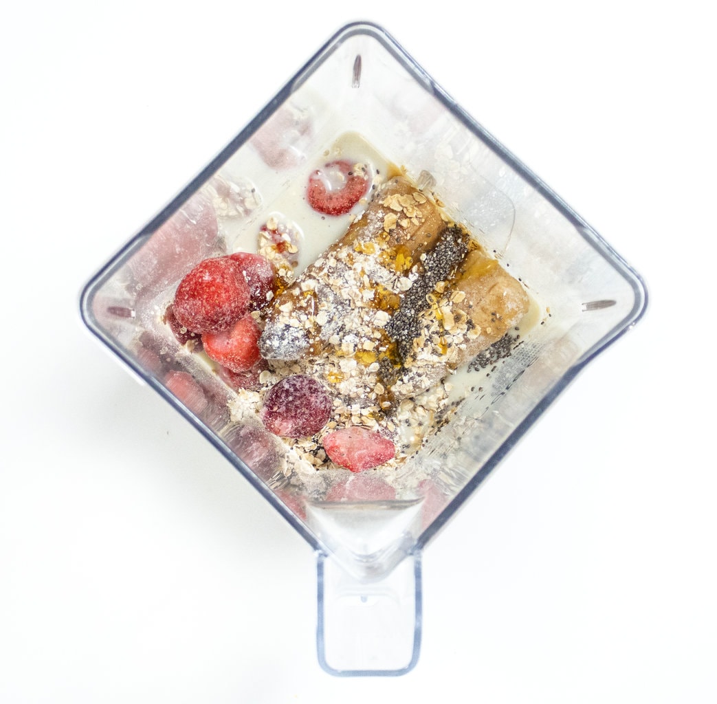 Clear blender full of ingredients for strawberry smoothie sitting on a white counter.