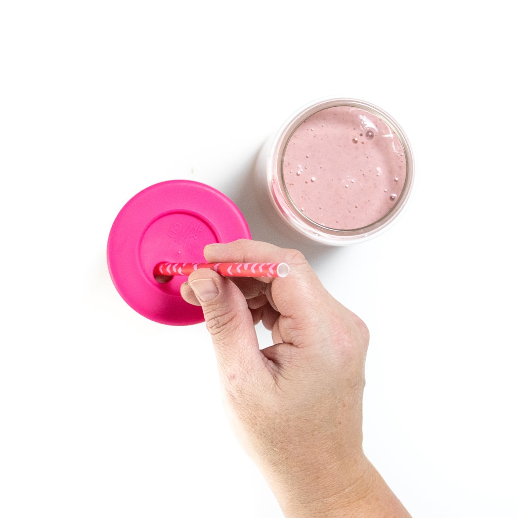 Hand placing a pink straw inside a kids glass glass full of strawberry smoothie with another glass sitting next to it.