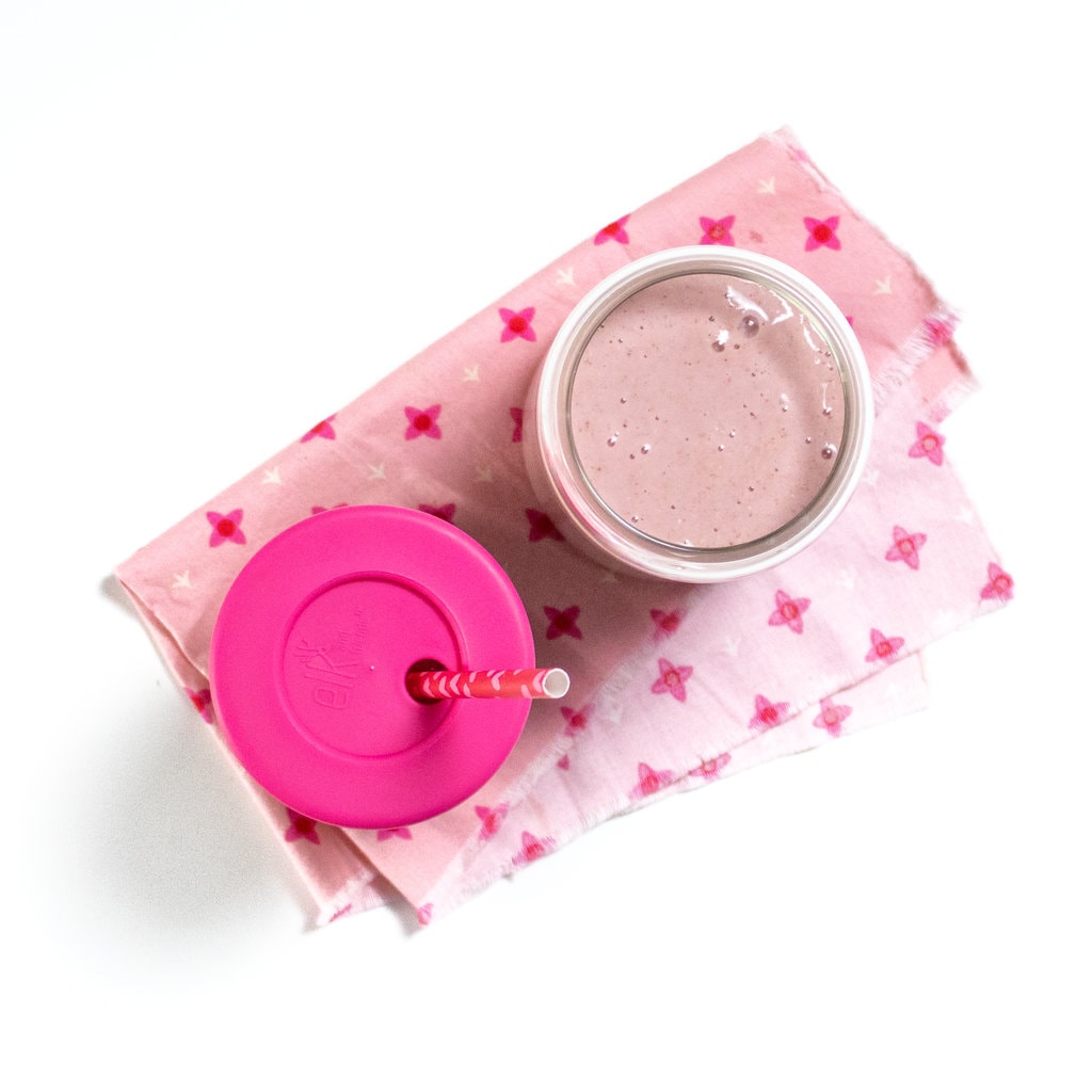 Two glasses, one with a pink lid, sitting on a pink napkin on a white counter.