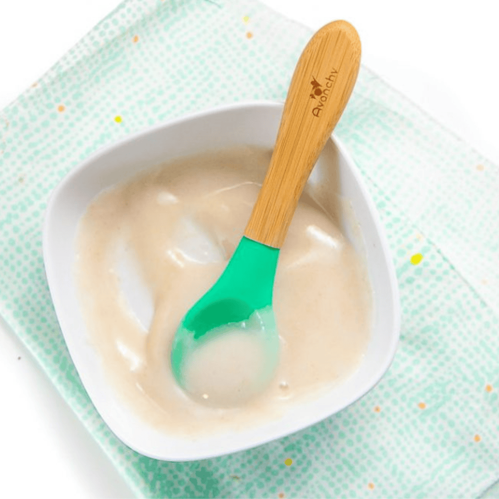 small white baby bowl filled with a smooth rice cereal for baby, sitting on a teal napkin with a teal spoon resting on top. 