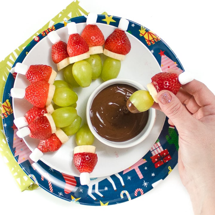Round plate with grinch fruit kabobs in a circle with small bowl of chocolate dipping sauce.