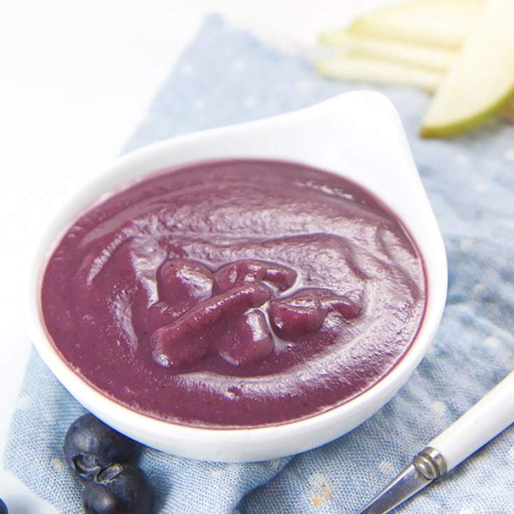 small white bowl on blue napkin with pears and blueberries scattered around. In the bowl is a smooth purple baby food purer.