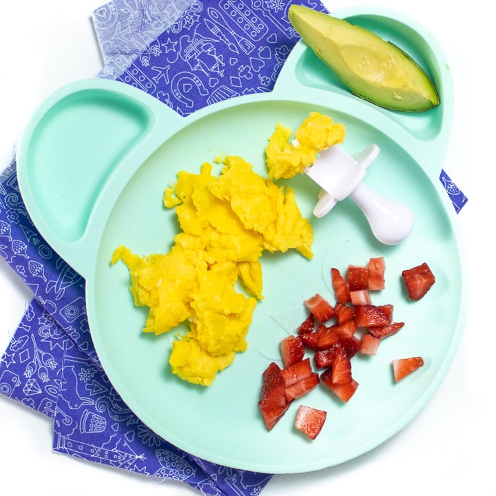 Teal plate on purple napkin with easy scrambled eggs, avocado and chopped strawberries, all setting on a white kitchen counter. 