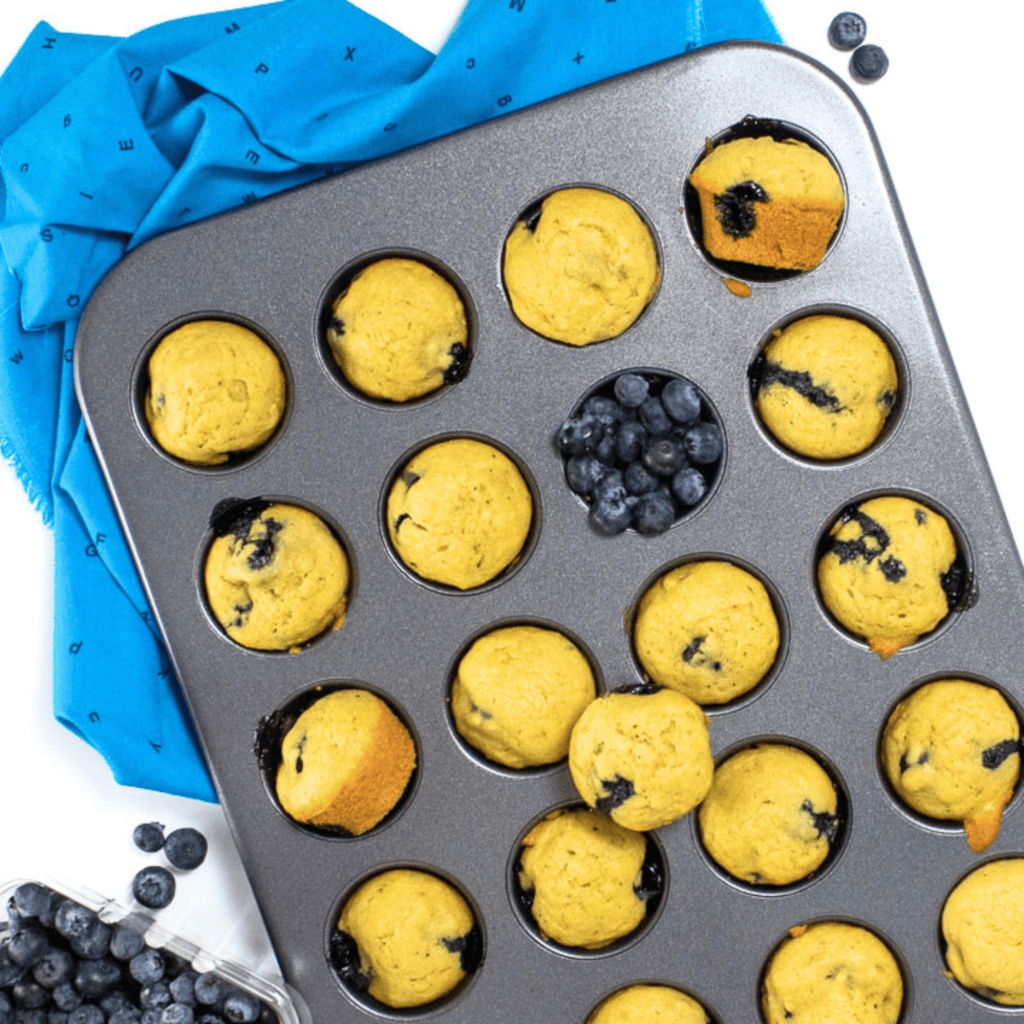 Muffin tin resting on a blue napkin filled with avocado blueberry muffins with blueberries scattered about. 
