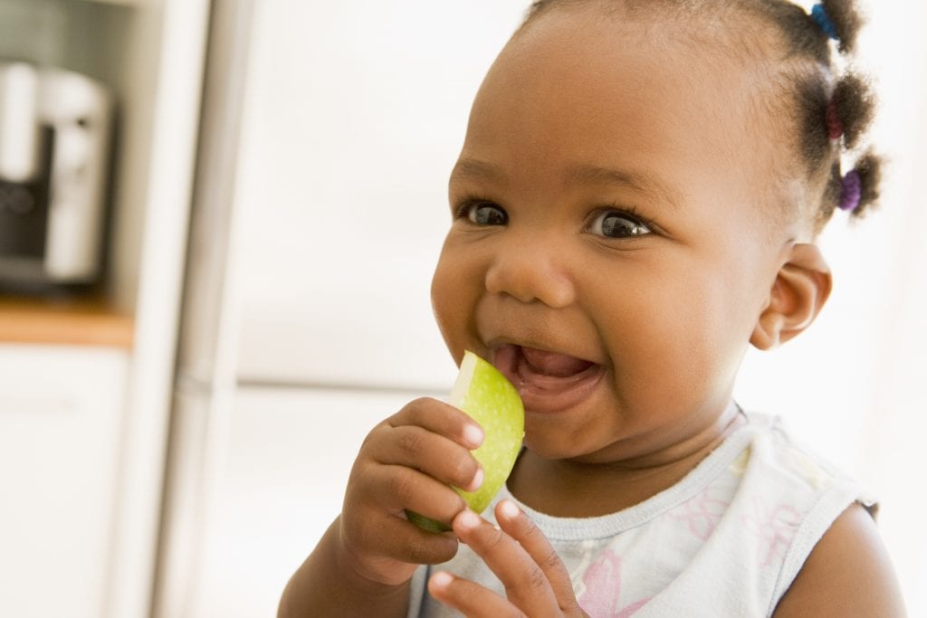 Baby girls with a bib feeding herself a piece of apple.