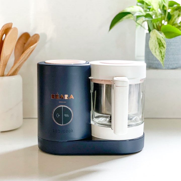 Beaba babycook on a white counter with a plant and white jar with wooden spoons sitting next to it.