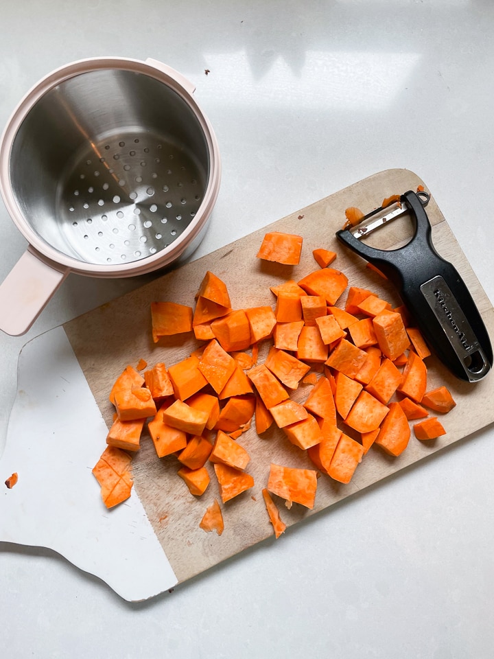 Wooden cutting board with chopped sweet potatoes a veggie peeler and the Beaba stainless steel basket sitting on a white counter.