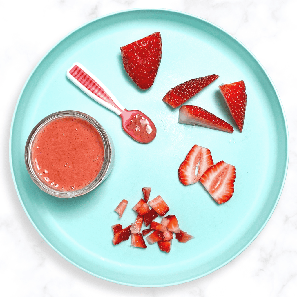 Teal baby plate with several different ways to serve strawberries to your baby. The plate is sitting on a marble countertop. 